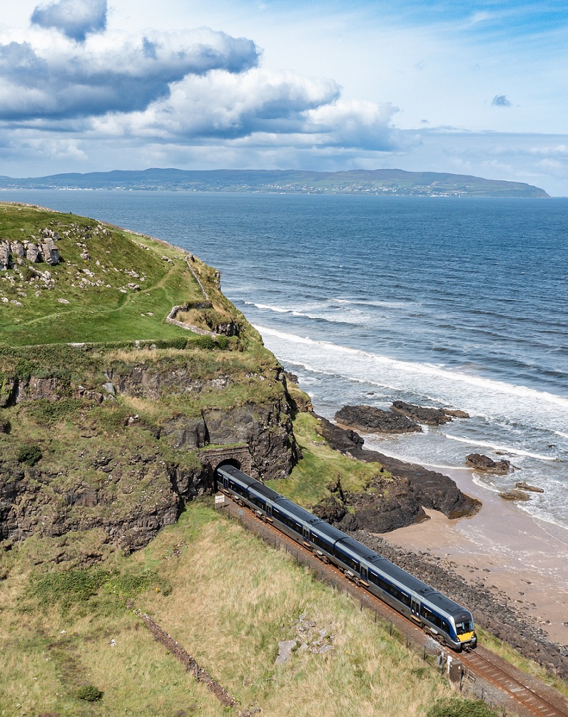 A photo of an NI Railways train passing along the coast on a bright, sunny day