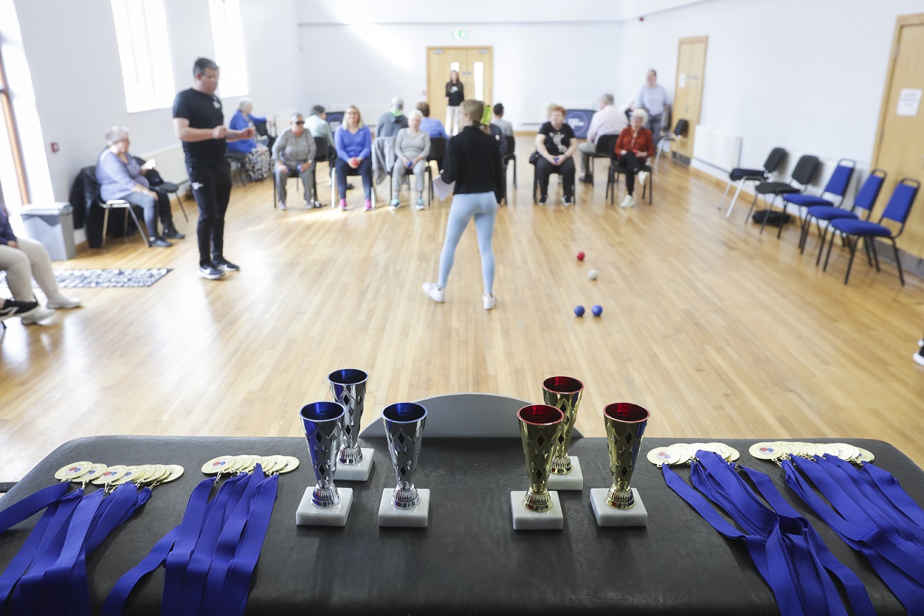 A photograph showing medals and trophies sitting on a table with people talking in the background